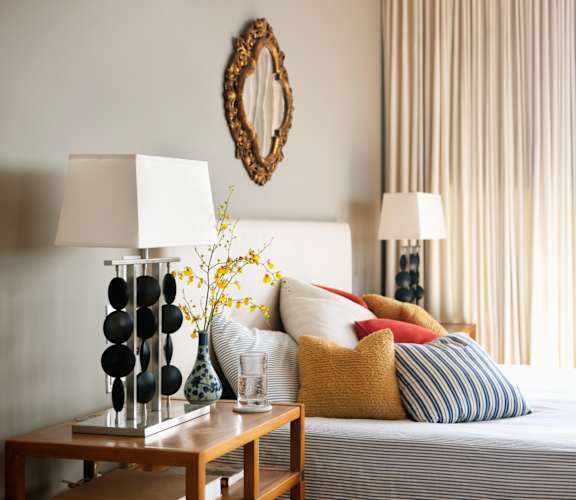 A bedroom with a bed, a nightstand with a lamp, a vase with flowers, and a mirror on the wall.