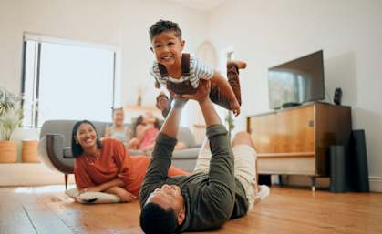 A man is lying on the floor with a child on his feet and another person sitting on the floor behind them.