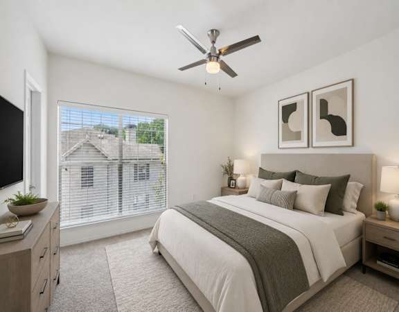 A bedroom with a large bed, a ceiling fan, and a window overlooking a house.