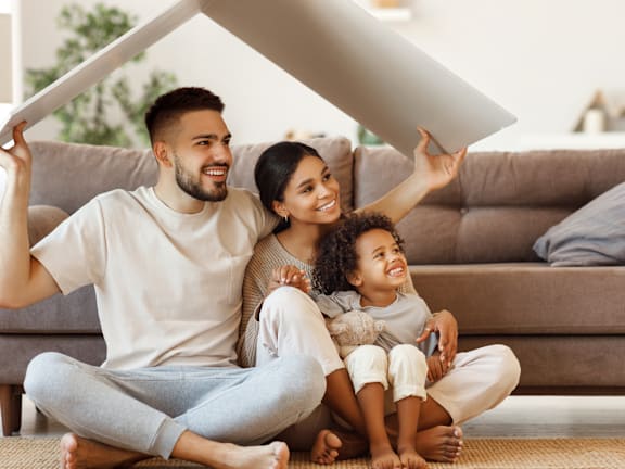 A family of three is sitting on the floor, holding a large piece of furniture.