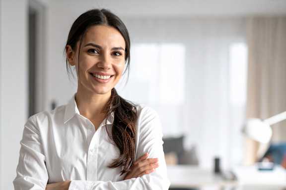 A woman in a white shirt is standing with her arms crossed in an office setting.