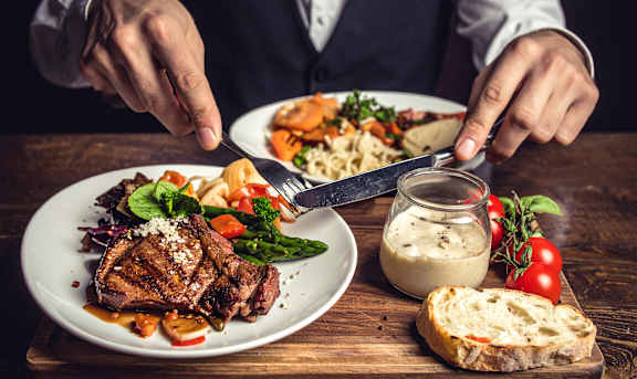 A person is cutting a steak on a plate with vegetables and bread.