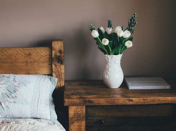 A vase of white flowers sits on a wooden table next to a pillow and a book.