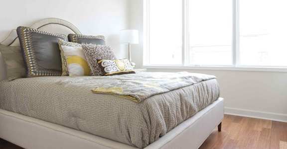 A bed with a grey comforter and a yellow pillow is in front of a window.
