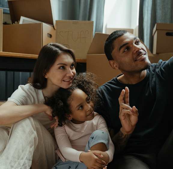 A family of three is sitting on the floor surrounded by moving boxes.