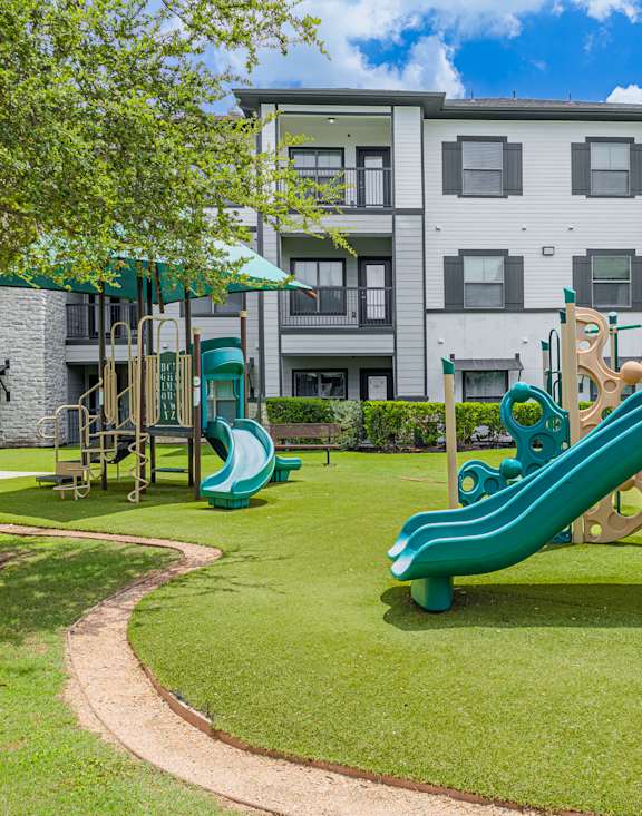 A playground with a green canopy and slide in the foreground and apartment buildings in the background.