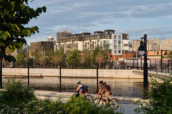 Two people riding bicycles on a path near a body of water with buildings in the background.