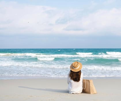 A woman in a straw hat sits on the beach looking out at the ocean.