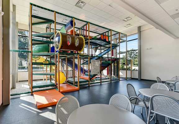 A playground structure with slides and climbing bars is inside a room with tables and chairs.