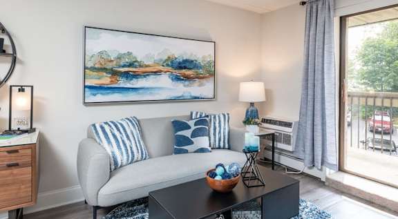 A living room with a grey couch, a painting on the wall, and a view of a balcony through the sliding glass door.