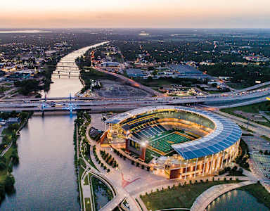 A large stadium with a green field is surrounded by a river and a bridge.