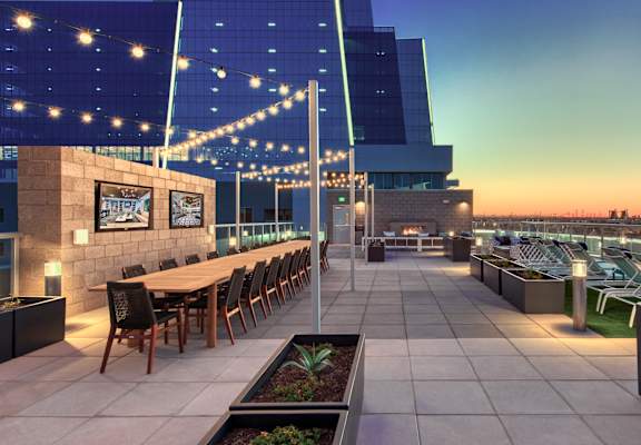 A rooftop patio with a long table and chairs, string lights, and a view of the city skyline at dusk.