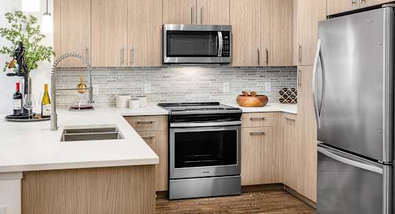 Modern model kitchen with light wood cabinetry, white quartz countertops, stainless steel appliances, a built-in microwave, and a peninsula with an undermount sink and bar seating.