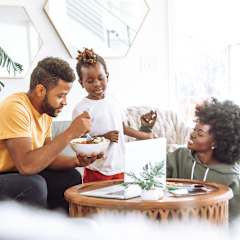 a man and woman sitting on a couch eating a bowl of food with a young girl standing