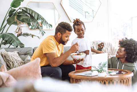 a man and woman sitting on a couch eating a bowl of food with a young girl standing