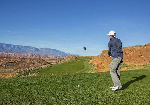 A man in a blue jacket and white cap is playing golf on a green field with mountains in the background.