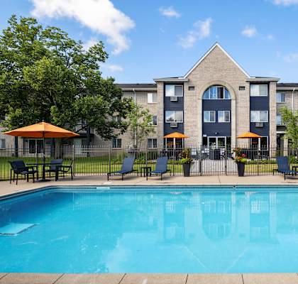 A swimming pool in front of a building with a patio and chairs.