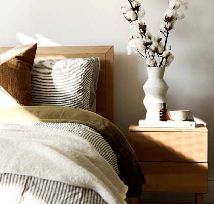 A bed with a white pillow and a brown pillow with a tan pillow case with a white vase with white flowers on a nightstand.