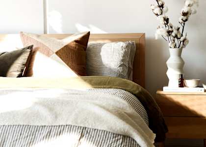 A bed with a white pillow and a brown pillow with a tan pillow case with a white vase with white flowers on a nightstand.