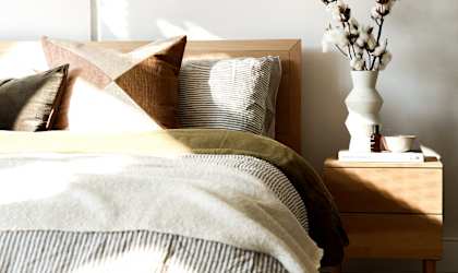 A bed with a white pillow and a brown pillow with a tan pillow case with a white vase with white flowers on a nightstand.