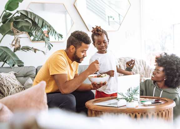 a man and woman sitting on a couch eating a bowl of food with a young girl standing