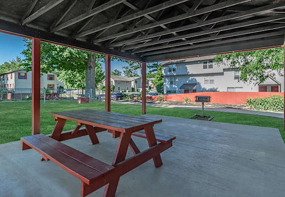 A picnic table under a covered area with a grassy area and buildings in the background.