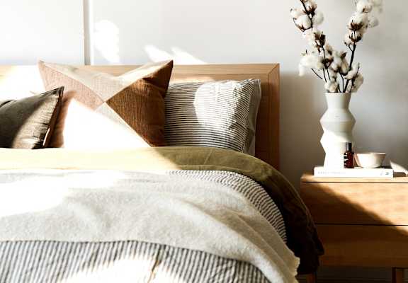 A bed with a white pillow and a brown pillow with a tan pillow case with a white vase with white flowers on a nightstand.