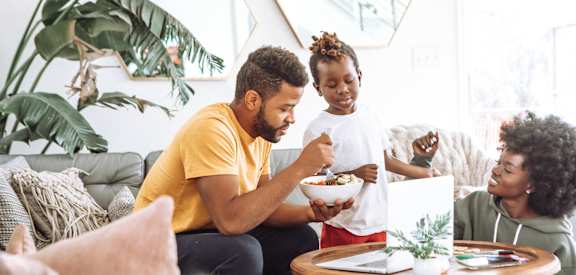 a man and woman sitting on a couch eating a bowl of food with a young girl standing