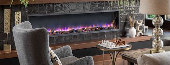A grey chair is in front of a fireplace with a chess board on the table.