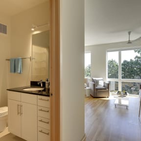 Glimpse into Flat B studio apartment floor plan at Coze Flats, bathroom has black granite counter, white cabinets. Past the bathroom is the living and sleeping area with oversized, East facing windows with treetop view