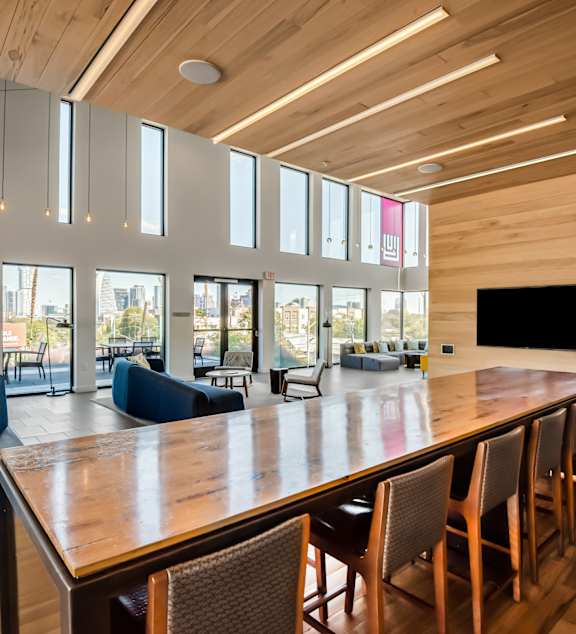 A long wooden table is surrounded by chairs in a room with a high ceiling and large windows.
