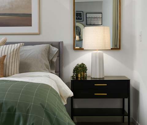 A bedroom with a bed, a nightstand with a lamp and a mirror above it.