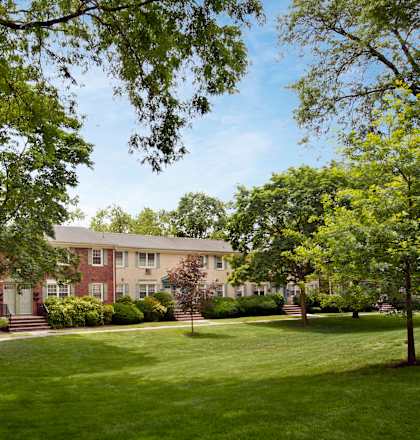 A large house with a red brick exterior is surrounded by a well-manicured lawn and trees at Rivercrest Apartments, Piscataway , New Jersey