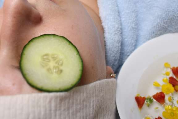 A person with a cucumber slice on their nose and a bowl of flowers nearby.