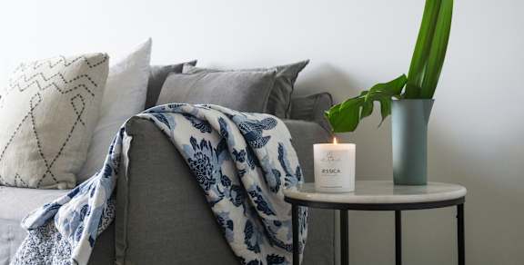 A grey couch with a blue and white blanket draped over it, a lit candle, and a green plant on a small table.