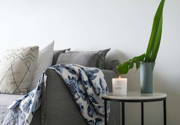 A grey couch with a blue and white blanket draped over it, a lit candle, and a green plant on a small table.