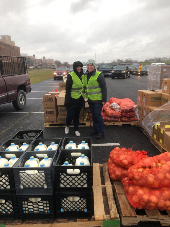two women in yellow vests stand in a parking lot with crates of potatoes and bags of