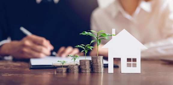 stacks of coins on a desk with a cutout of a house and plants
