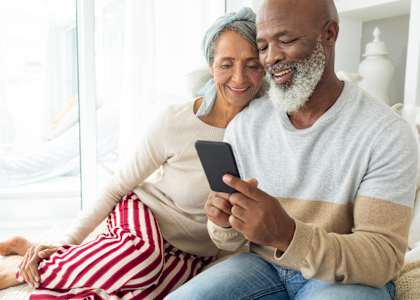 An older couple is sitting on a couch, with the man holding a smartphone.