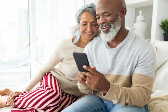 An older couple is sitting on a couch, with the man holding a smartphone.