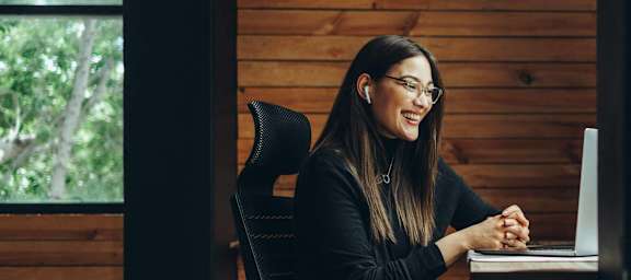 A woman is sitting at a desk with a laptop, wearing glasses and a black top.