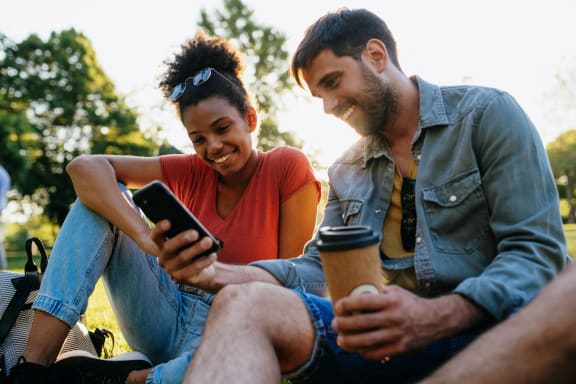 a man and a woman sitting on the grass looking at a cell phone