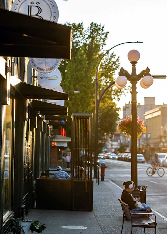 Sidewalk with storefronts and a person sitting on a bench.