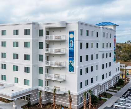 A large white hotel with a blue roof and a parking lot in front.