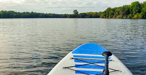 A blue paddleboard is in the foreground of a calm lake with trees in the background.