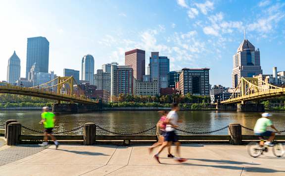 Three people jogging on a sidewalk with a city skyline in the background.