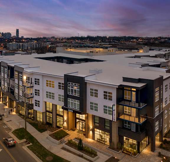 A modern apartment complex is lit up at dusk with cars driving on the road in front.