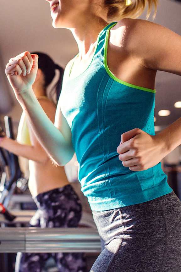 A woman in a blue tank top and grey leggings is running on a treadmill in a gym.