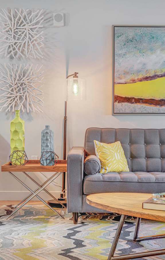 A grey couch with a yellow pillow sits in front of a painting in a living room.