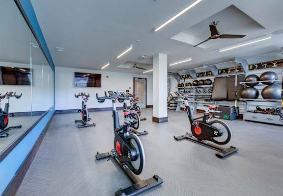 A gym with a row of stationary bikes and a fan on the ceiling.
